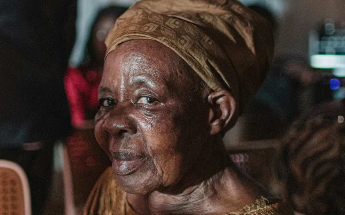 An older woman in a gold headwrap and embroidered dress, smiling softly.