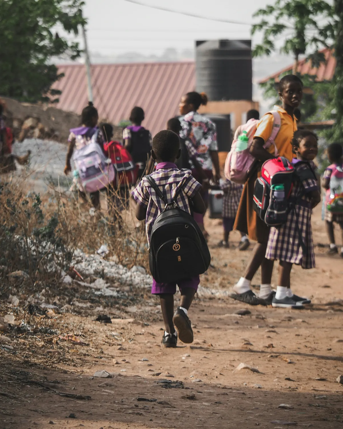 Children in checked school uniforms walk along a dirt road with their school bags; one girl looks back and smiles.
